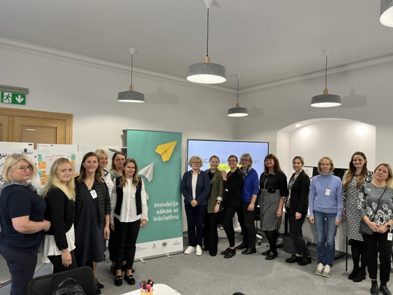 Dalībnieku grupa — aptuveni piecpadsmit cilvēki — pozē fotogrāfijai pie programmas reklāmkaroga.
